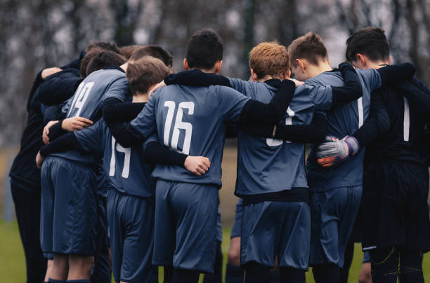 Soccer team with junior coach gathering together in a circle, to strategise and motivate. Football youth team standing together outdooe on grass pitch